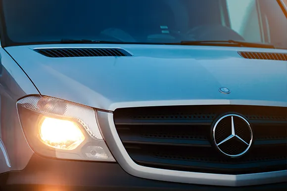 The front of a Mercedes-Benz Sprinter with its headlights on.