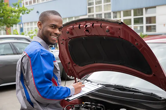 A young auto mechanic in front of an open car hood while writing on a list on his clipboard